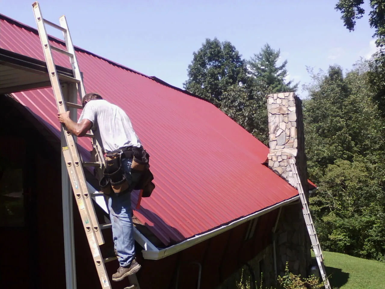 Roofer on a ladder beside a red metal roof and stone chimney — Soffit & Fascia Repair by Roofing Brothers in Hartsel